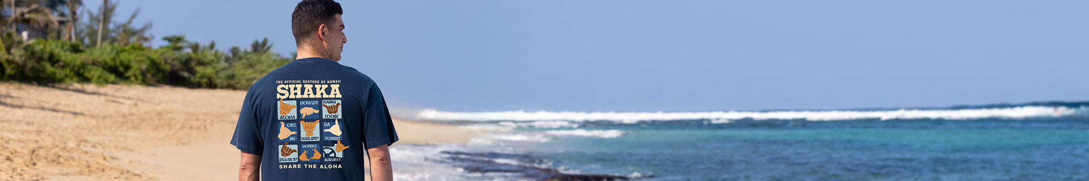 A man stands on a sandy beach facing the ocean, wearing a dark navy blue t-shirt with a colorful graphic and 'SHARKA' printed on the back.