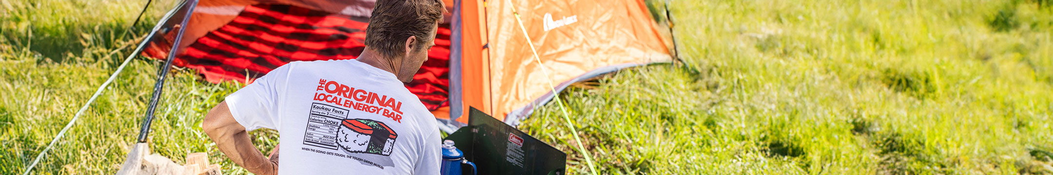 A man wearing a white t-shirt with a red and black graphic print stands next to an orange tent in a grassy outdoor setting.
