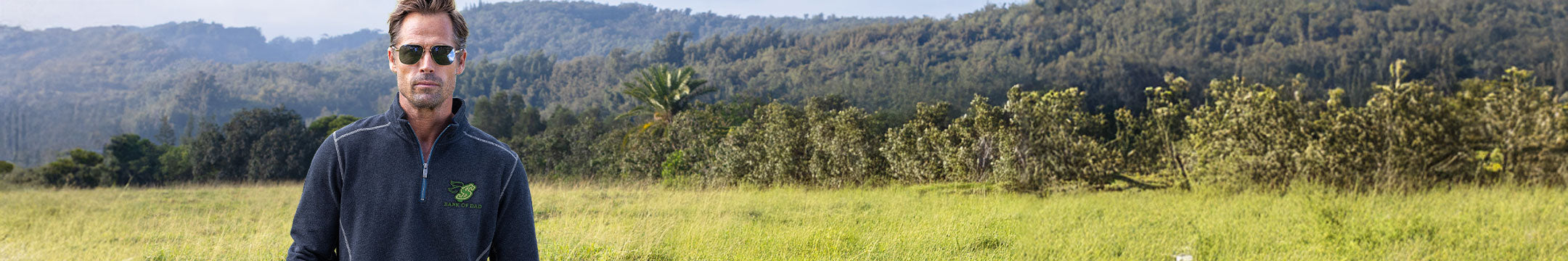 A man wearing a dark navy blue quarter-zip pullover and sunglasses stands in a grassy field with trees and hills in the background.