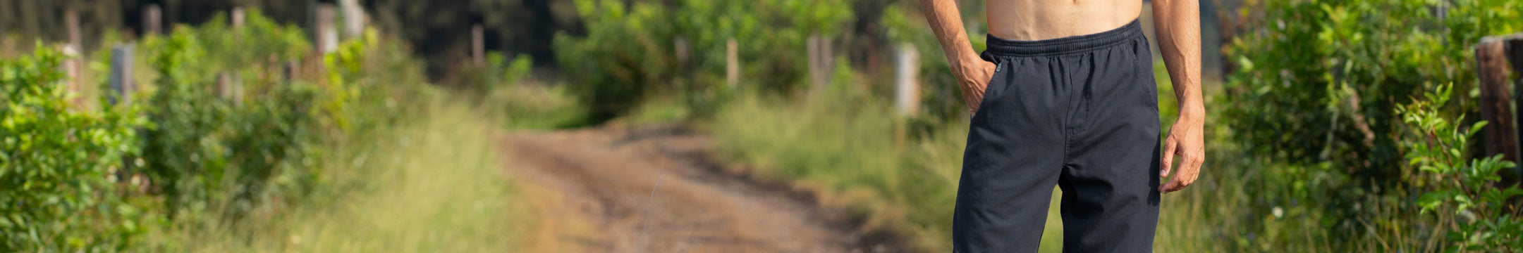A man wearing dark gray pants stands on a dirt path surrounded by green foliage, with his torso bare and his other arm in his pocket.