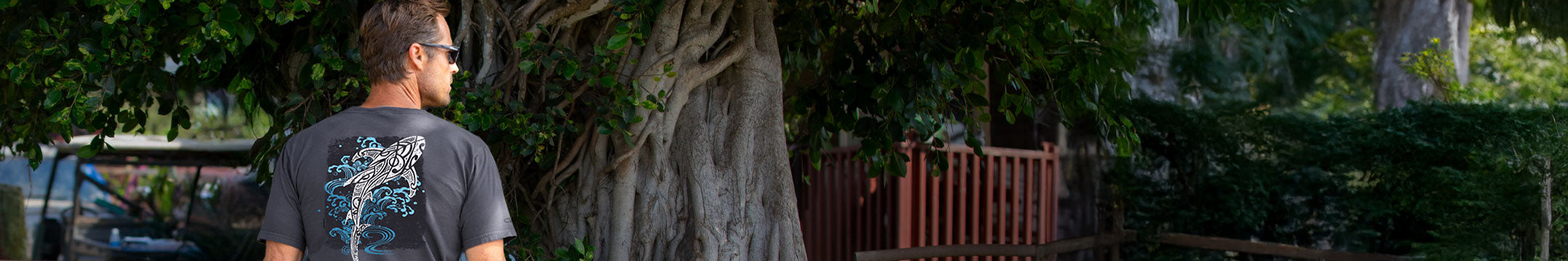 A man wearing a dark gray t-shirt with a blue and white shark graphic, standing outdoors in front of a large tree trunk.
