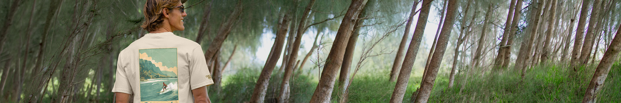 A man wearing a white t-shirt with a large graphic print of a surfer in the ocean on the back, standing in a forest of tall, slender trees.