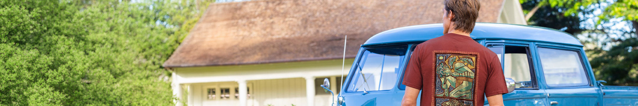 A man wearing a brown t-shirt with a colorful graphic print on the back stands in front of a blue vintage car and a house, viewed from behind.