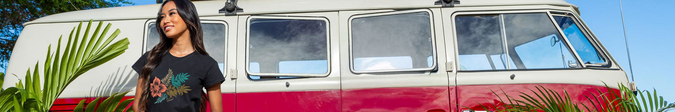 A woman wearing a black graphic t-shirt stands next to a vintage red and white Volkswagen camper van outdoors with palm leaves in the foreground.