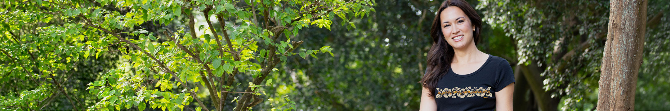 A woman with long dark hair stands outdoors in front of a lush green tree, wearing a black short-sleeve t-shirt with golden flower graphic print on the front.
