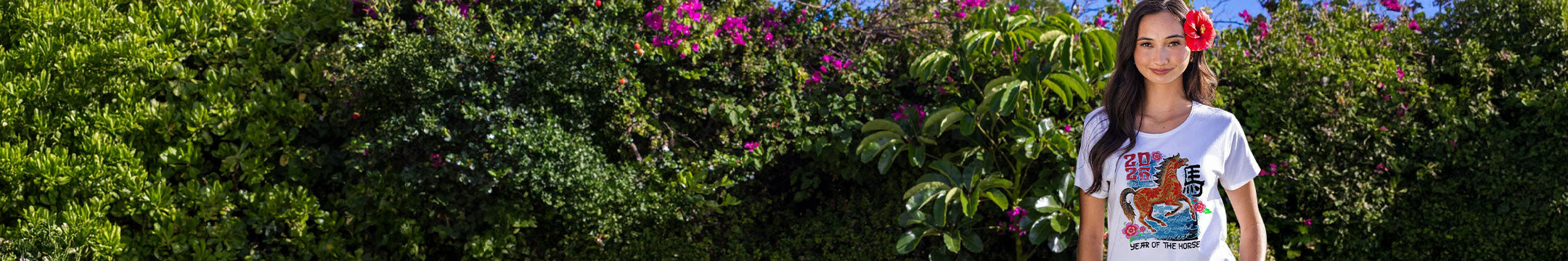 A woman wearing a white graphic t-shirt with a colorful cartoon horse print stands outdoors in front of a lush green hedge with pink flowers. She has a red flower in her hair.