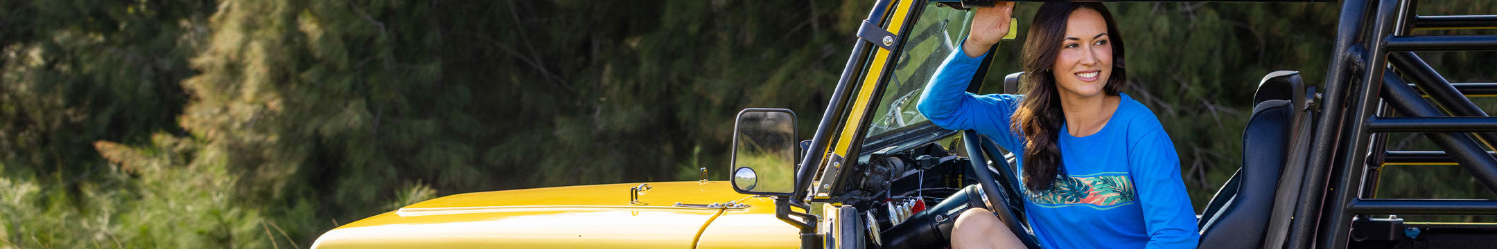 A woman smiling from inside a yellow off-road vehicle, with trees in the background. She is wearing a bright blue long-sleeve shirt with a colorful floral graphic band on front.