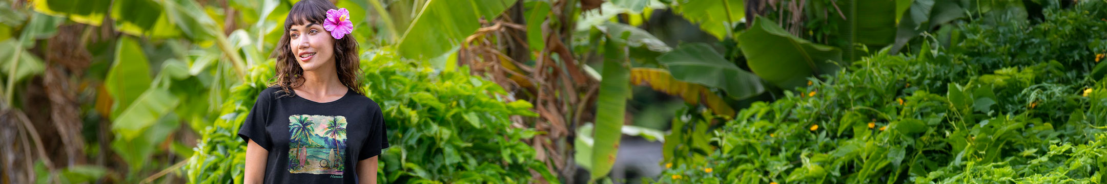 A woman wearing a black graphic t-shirt with a tropical beach print and a pink flower in her hair, standing in a lush green outdoor setting with banana leaves.