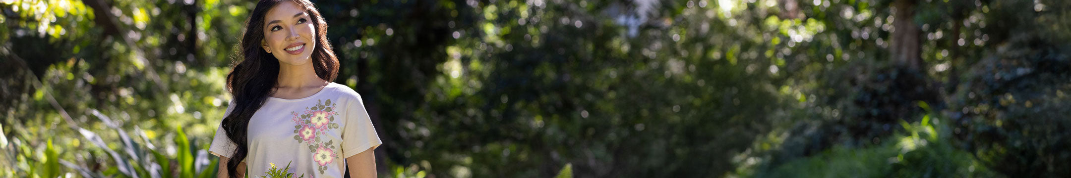 A woman standing outdoors in a sunlit forest, wearing a light-colored beige short-sleeve top with a floral print. The background is heavily blurred green foliage.