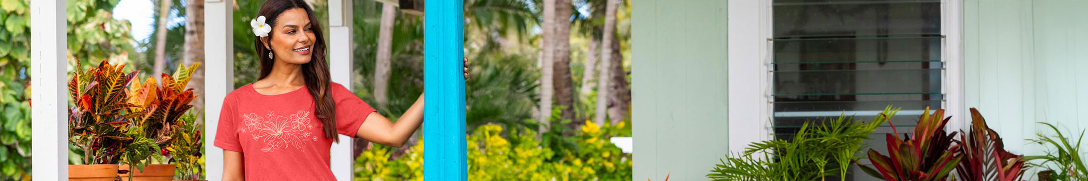 A woman wearing a red short-sleeve top stands outdoors in front of a white house with tropical plants and palm trees visible in the background.