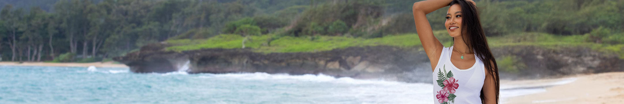 A woman stands on a sandy beach wearing a white tank top with a colorful floral graphic on the front, with ocean waves and a green forested coastline in the background.