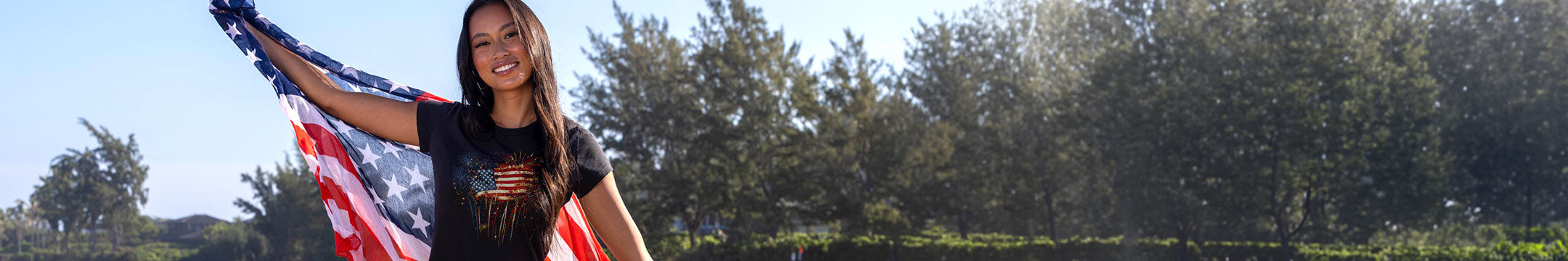 A woman outdoors holding a large American flag, wearing a black graphic t-shirt. The background features a line of green trees under a bright blue sky.