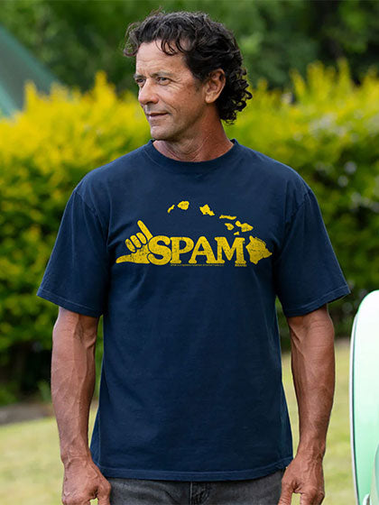A man wearing a navy blue t-shirt with a yellow 'SPAM' graphic and shaka hand sign on the front, standing outdoors in front of green and yellow foliage.