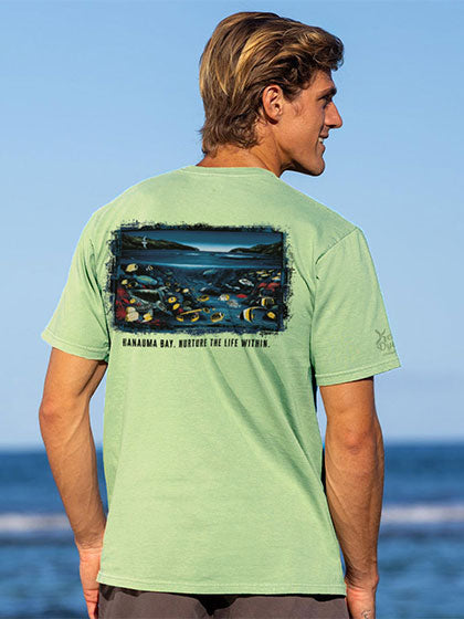 A man wearing a light green t-shirt with a large back graphic depicting the ocean and sea creatures below and the text 'HANAUMA BAY, NURTURE THE LIFE WITHIN', photographed outdoors against a blue sky and ocean background.