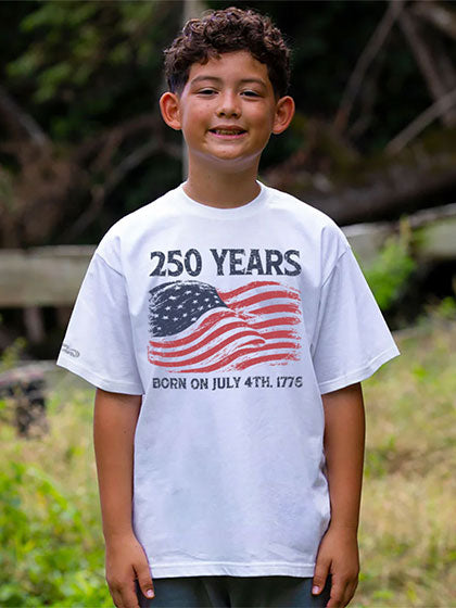 A smiling young boy wearing a white graphic t-shirt with a distressed American flag and the text '250 YEARS BORN ON JULY 4TH 1775' printed on the front, set against a blurred outdoor background.