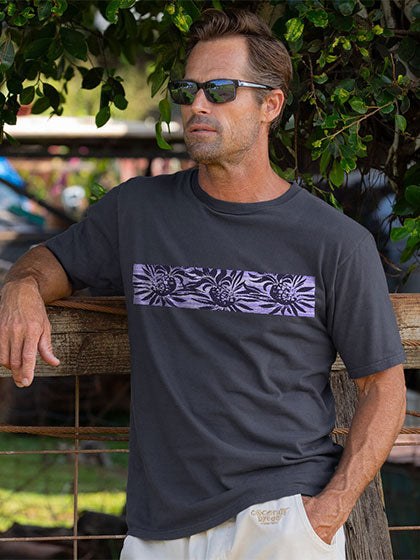 A man wearing a dark charcoal gray t-shirt with a horizontal purple and gray floral pattern across the chest, leaning on a wooden fence outdoors.