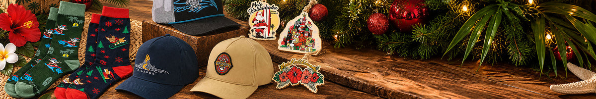 A festive holiday-themed product display featuring Christmas ornaments, patterned socks, baseball caps, and a small figurine arranged on a rustic wooden surface next to evergreen branches with warm string lights.