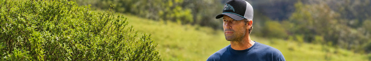 A man wearing a navy blue t-shirt and a black and gray baseball cap stands outdoors in a grassy field with trees in the background.