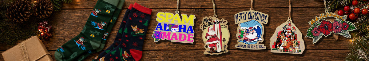 A row of colorful Christmas-themed hanging ornaments and stockings displayed on a rustic wooden surface, with pinecones, evergreen branches, and a wrapped gift partially visible around the edges.