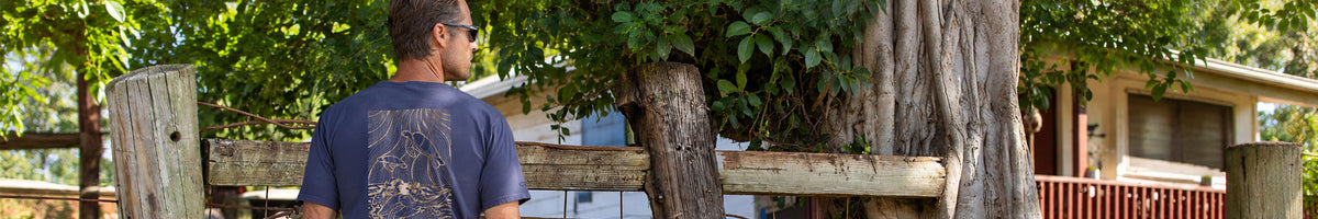 A man wearing a navy blue t-shirt with a gold graphic design on the back, standing outdoors in front of a rustic wooden fence and trees.