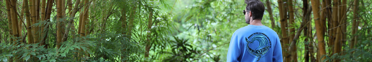 A man viewed from behind wearing a light blue crewneck sweatshirt with a circular graphic on the back, standing in a dense green forest with bamboo.