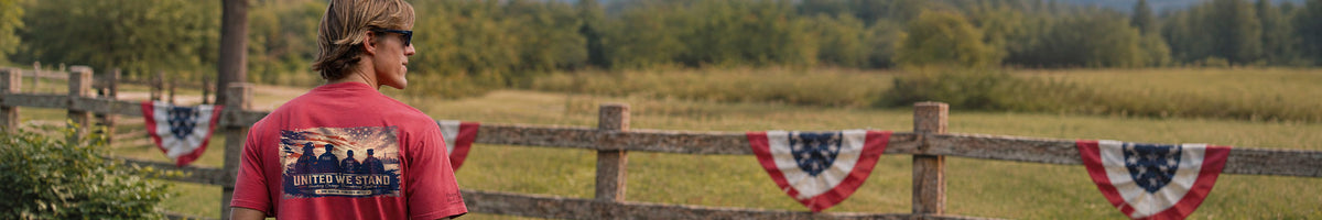 A man wearing a red t-shirt with a patriotic graphic print stands in front of a wooden fence decorated with American flag bunting in a field.