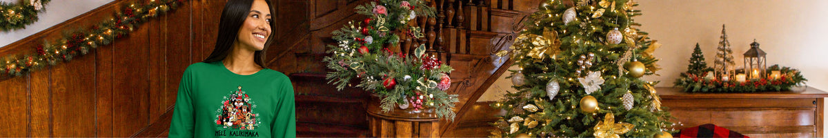 A woman wearing a festive green Christmas long sleeve with a holiday graphic stands in a decorated home interior next to a lit Christmas tree and staircase.