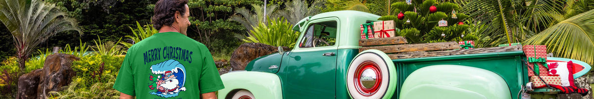 A man wearing a green t-shirt with a 'Merry Christmas' graphic stands next to a vintage green and white pickup truck loaded with Christmas decorations and wrapped gifts in a tropical outdoor setting.