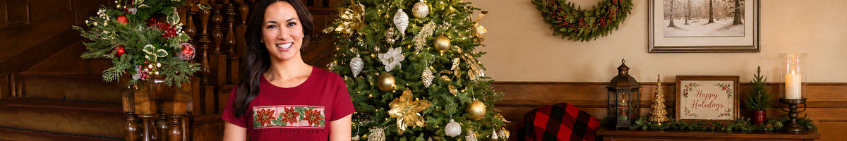 A woman wearing a festive red Christmas short sleeve with a colorful holiday pattern across the chest, standing in a decorated living room with a Christmas tree, wreath, and fireplace.