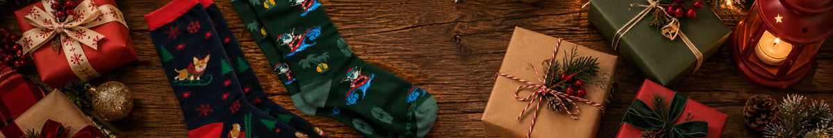 A festive flat-lay of Christmas socks on a rustic wooden table, surrounded by wrapped gift boxes, ornaments, pinecones, and a lit candle.