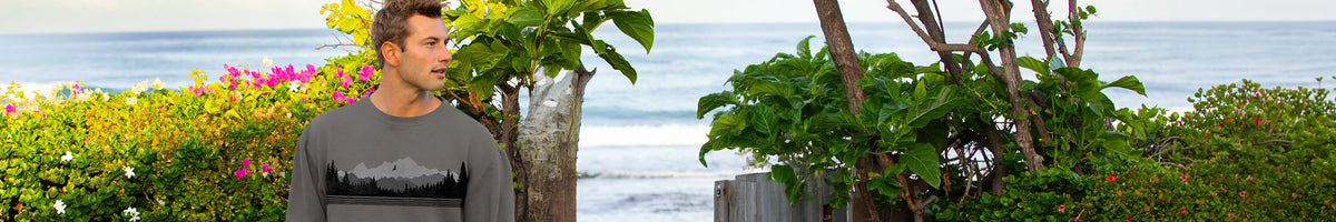 A man wearing a dark gray long-sleeve t-shirt with a black graphic print, standing outdoors in front of tropical green foliage and a scenic ocean background.