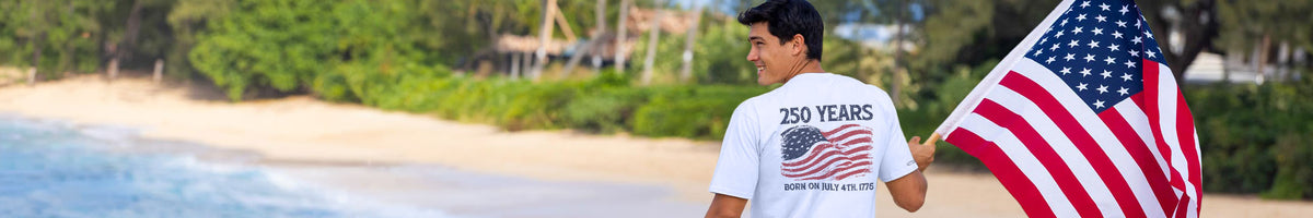 A man on a beach wearing a white t-shirt with a patriotic 250th anniversary graphic on the back, holding a large American flag.