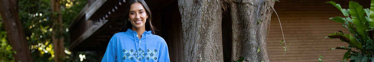 A woman wearing a bright blue embroidered tunic stands outdoors in front of a large tree trunk, with a wooden structure and green foliage in the background.