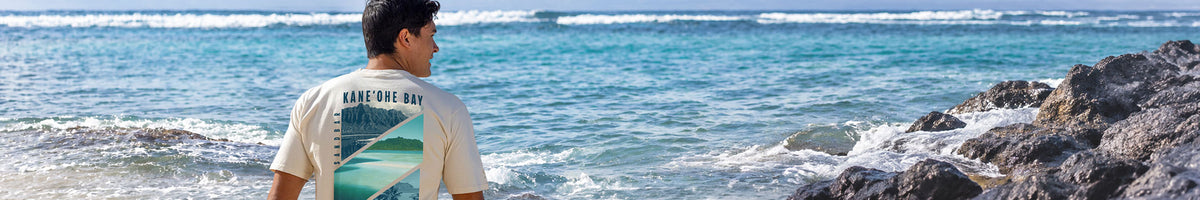 A man wearing a cream-colored t-shirt with a graphic print stands on a rocky beach looking out towards the ocean.