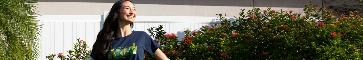 A woman smiling outdoors in front of a white fence and green bushes with red flowers, wearing a navy blue graphic t-shirt.