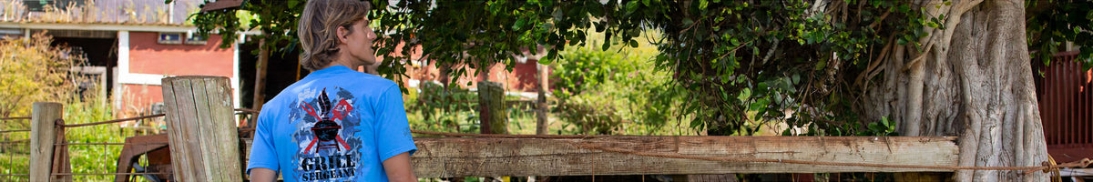 A man wearing a bright blue t-shirt with a black graphic of a rabbit standing in front of a rustic wooden fence and a large tree outdoors.