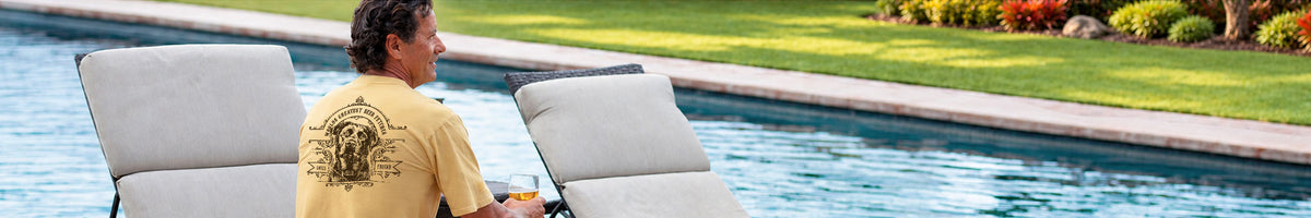 A man wearing a yellow graphic t-shirt sits on a white outdoor poolside lounge chair next to a swimming pool with a green lawn in the background.