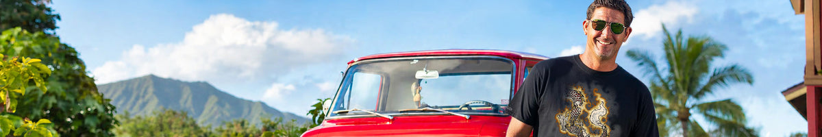 A man wearing a black graphic t-shirt and sunglasses stands in front of a vintage red pickup truck outdoors with mountains, palm trees, and a blue sky in the background.