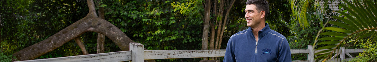 A man wearing a navy blue quarter-zip pullover shirt stands outdoors in front of a wooden fence and lush green foliage.
