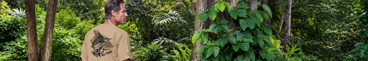 A man wearing a tan t-shirt with a black graphic print on the back, standing in a lush green forest.