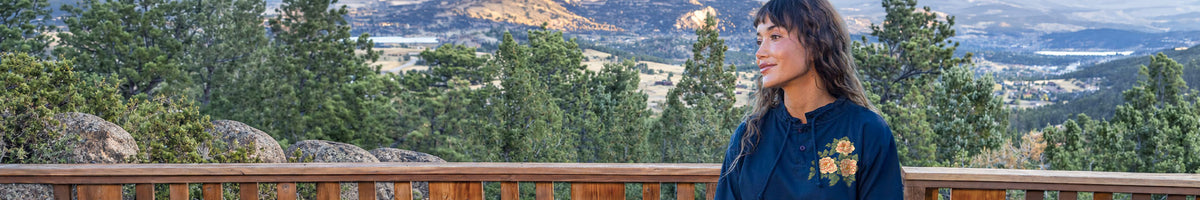 A woman with dark curly hair wearing a navy blue embroidered shirt stands on a wooden deck overlooking a scenic mountain landscape with pine trees.