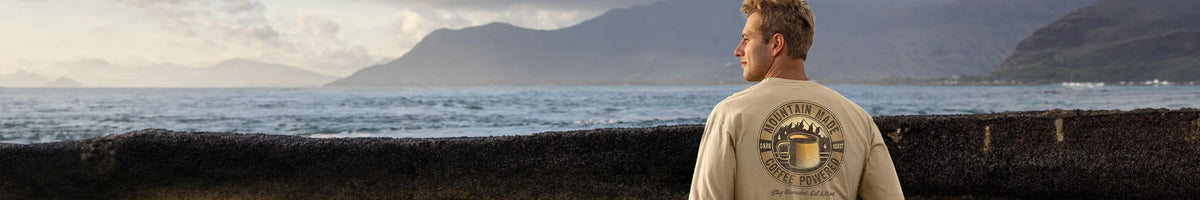 A man stands on a rocky shoreline with the ocean and mountains in the background, wearing a light beige T-shirt with a large circular graphic on the back featuring a beer mug.