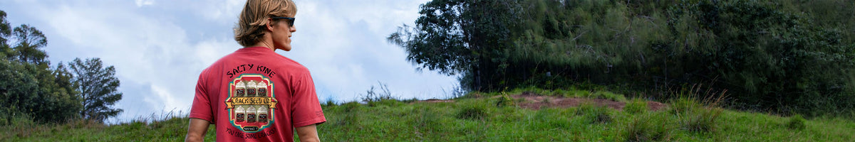 A man wearing a red t-shirt with a large graphic print on the back, standing outdoors on a grassy hill with trees and a cloudy sky in the background.