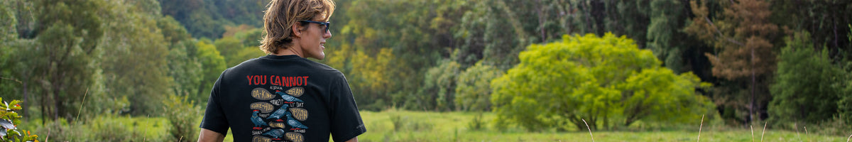 A man wearing a black graphic t-shirt with a red and blue design on the back, standing in a grassy field with trees in the background.