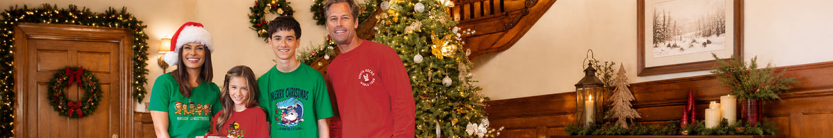 A family of four posing in a decorated holiday home. The mother wears a green  t-shirt and Santa hat, the father wears a green Christmas' t-shirt, the young boy wears a green t-shirt, and the older man wears a red long sleeve with a white logo.