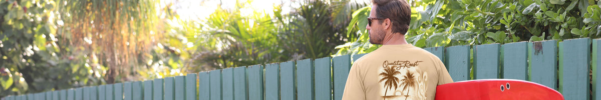 A man wearing a beige graphic t-shirt with palm tree designs on the back, standing outdoors next to a green fence and holding a red surfboard.