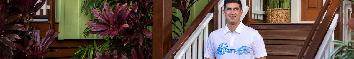 A man wearing a white polo shirt with a blue wave graphic stands on outdoor wooden stairs next to lush green tropical plants.