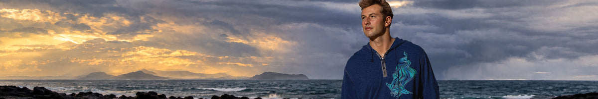 A man wearing a blue long-sleeve shirt with a teal graphic stands against a dramatic ocean sunset background.