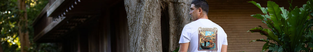 A man wearing a white t-shirt with a large graphic print on the back, standing outdoors next to a large tree trunk in a tropical setting.