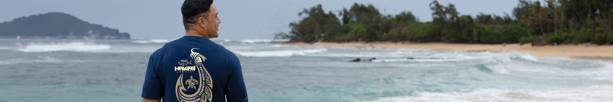 A man wearing a navy blue t-shirt with a gold graphic print stands with his back to the camera, looking out over a tropical beach with waves and trees.